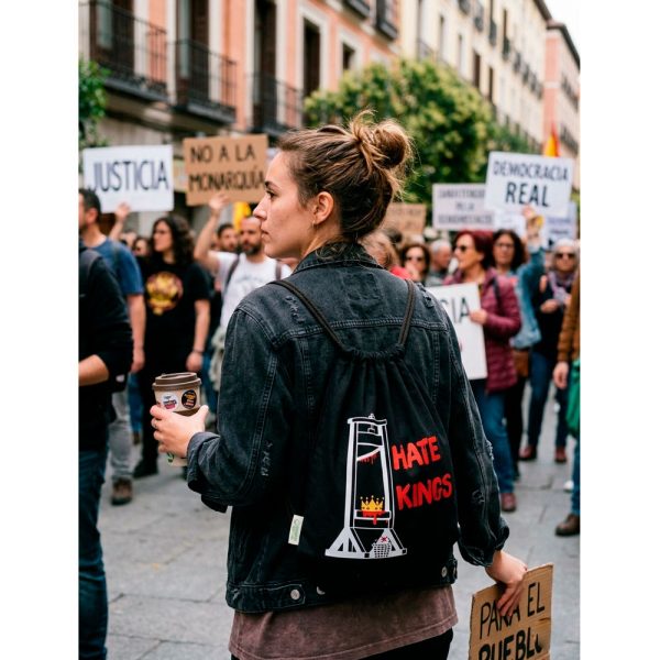 Mujer de espaldas con mochila Hate Kings de Ouh Rebel caminando en una protesta con carteles contra la monarquía.