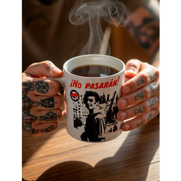Manos tatuadas sosteniendo una taza humeante de Ouh Rebel con el diseño "¡No Pasarán!" en un ambiente cálido.