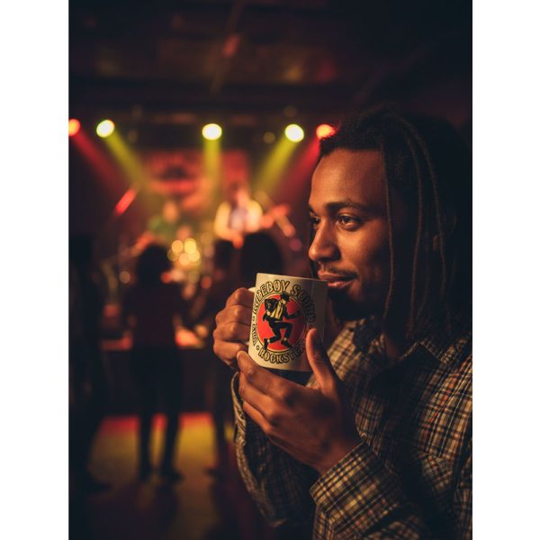 Un hombre con rastas y una camisa de cuadros sostiene la taza "Rudeboy Sound" de Ouh Rebel en un concierto, con luces de escenario de colores borrosas de fondo.